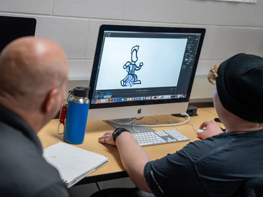 Student sitting at a desk next to an instructor animating something on a computer