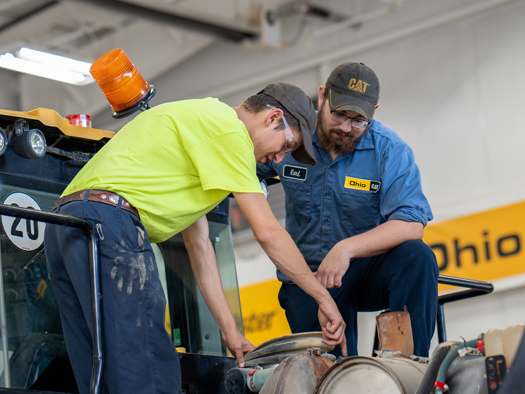 Two males working on an engine next to caterpillar equipment