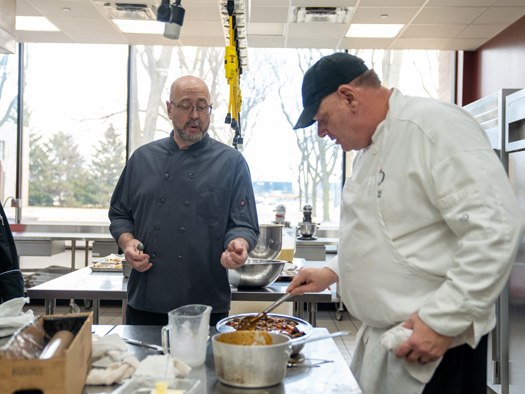 An instructor and another individual working in a kitchen together, with the instructor giving guidance to the other individual