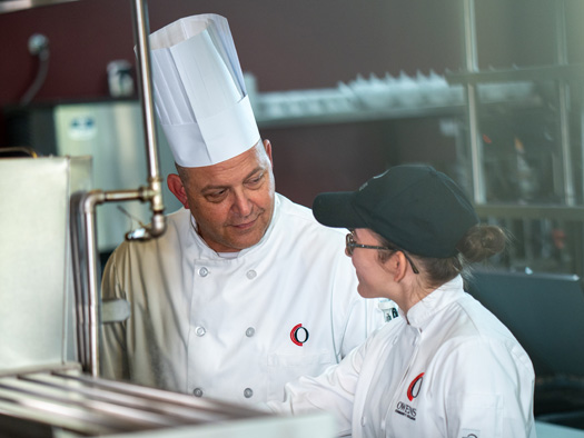 A chef standing next to another individual smiling and giving guidance in a kitchen