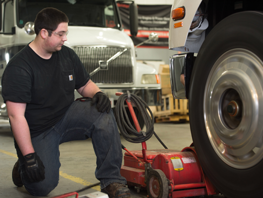 student kneeling down next to a large tire of a diesel truck
