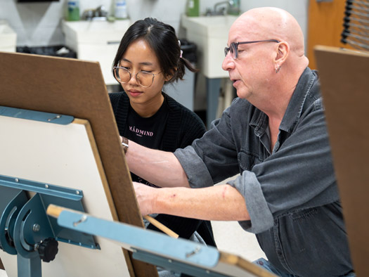 A teacher and a student in front of an easel, the teacher giving instruction/guidance to the student