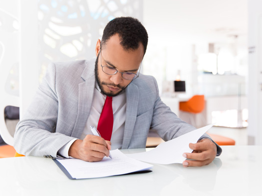 a man wearing a suit with a red tie sitting at a desk writing into a document