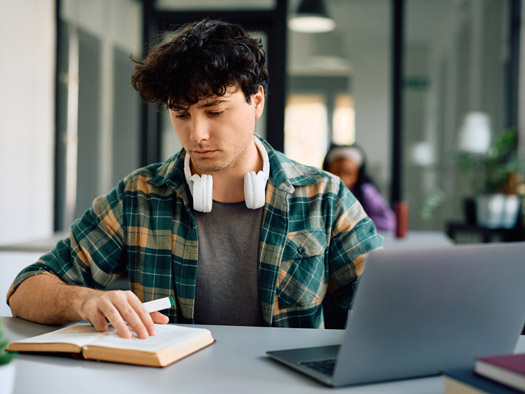 student sitting at desk taking notes with headphones around his neck
