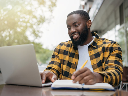student smiling while taking notes and looking at computer screen