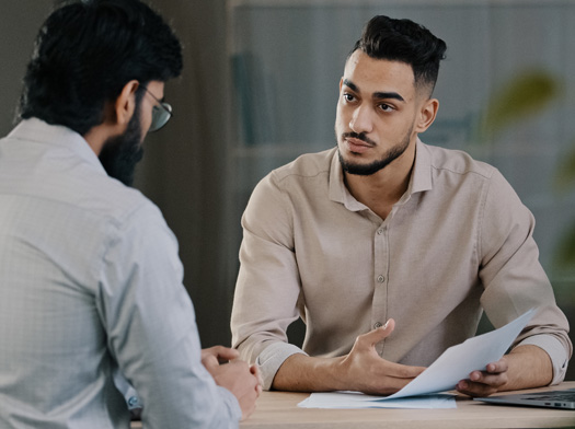 two guys sitting at a table discussing something while young man holding paper.