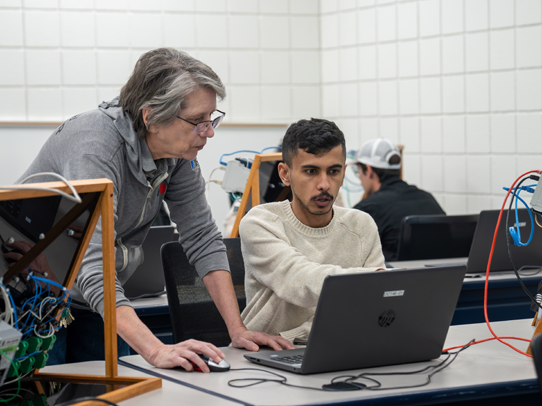 Male student sitting at a desk, with a female instructor standing close by looking at his laptop