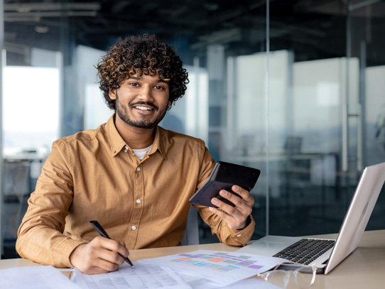 Man sitting at a desk in a corporate setting working on a laptop smiling at the camera