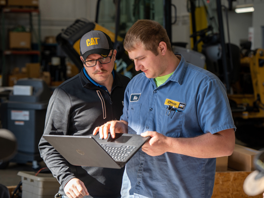 Two males standing next to each other looking at a laptop next to caterpillar equipment