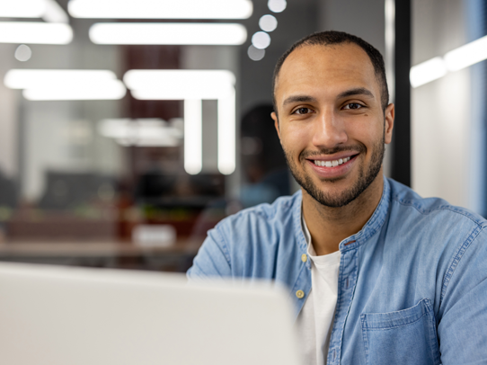 Male sitting in front of a laptop looking at the camera smiling